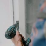 Crop bearded guy installing new wooden window with contemporary screwdriver and metal hinge during renovation process in apartment